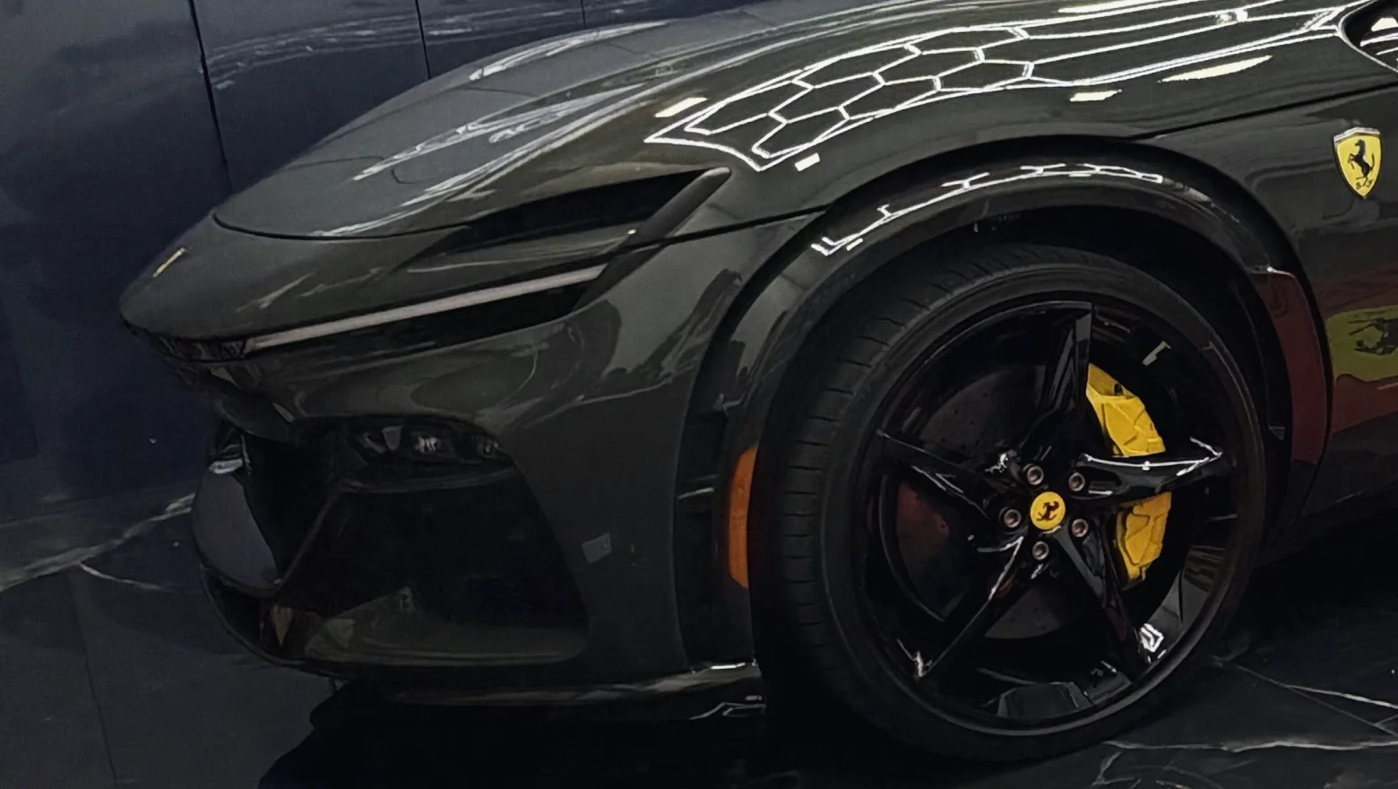 Front wheel and yellow brake caliper of a gray Ferrari Purosangue, hexagon lights reflected in the hood