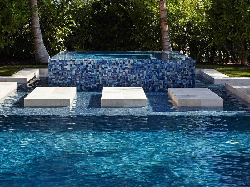 A raised spa clad in blue glass mosaic spilling into a swimming pool, with stone stepping pads