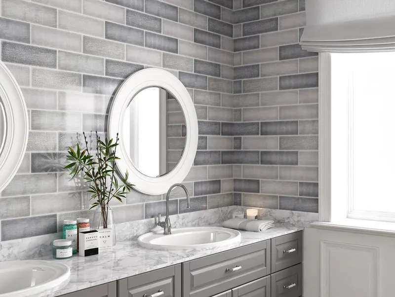 A bright bathroom vanity with grey textured subway tile on two walls and oval mirrors