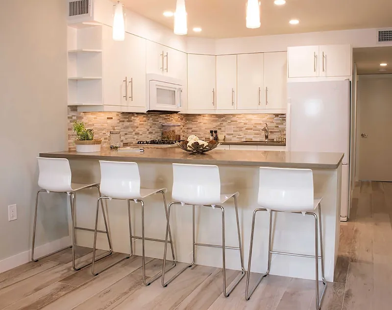 A bright white kitchen with a beige linear stone mosaic backsplash and a quartz peninsula