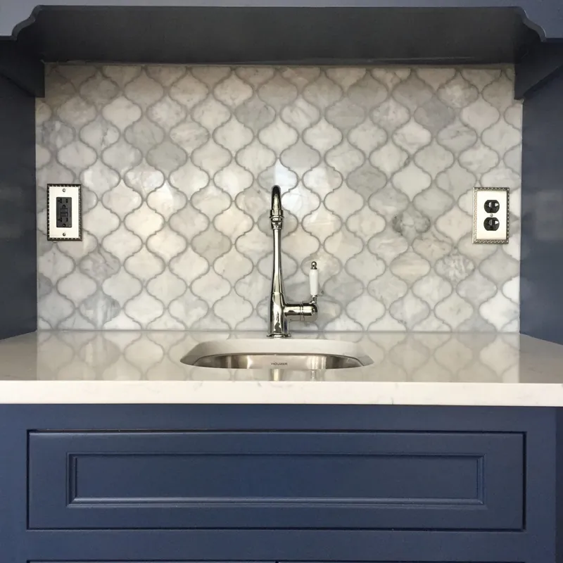 A marble arabesque mosaic backsplash behind a wet bar sink with navy cabinetry